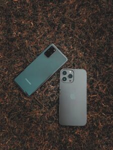 Two smartphones, one gray and one green, on a brown surface. The gray phone has a camera.