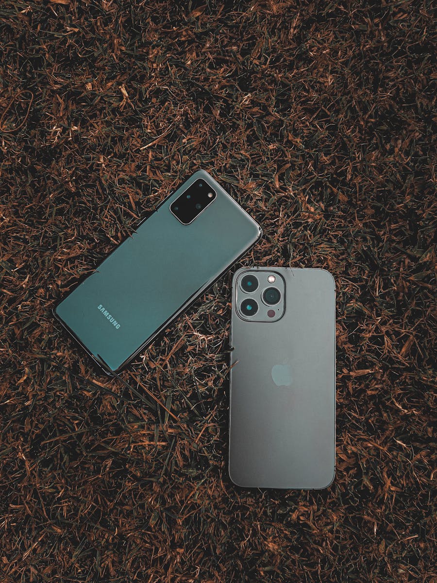 Two smartphones, one gray and one green, on a brown surface. The gray phone has a camera.